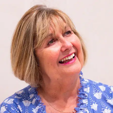 Janine looking off to the side in front of a plain background and wearing a blue frilled top with white flower patterns