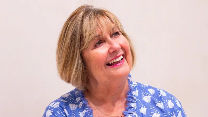 Janine looking off to the side in front of a plain background and wearing a blue frilled top with white flower patterns
