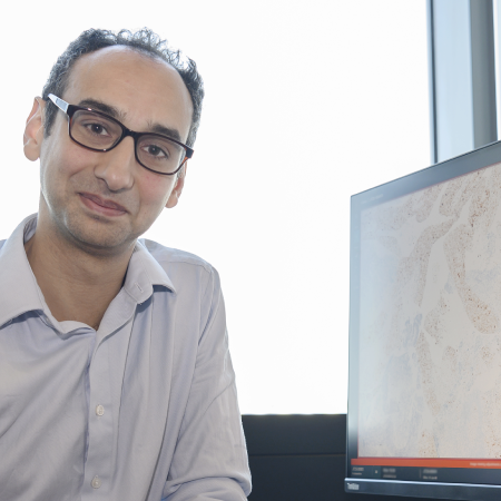John Tadross sitting at a desk with a window behind him. On the computer screen is a pathology image from a microscope showing stained cells.