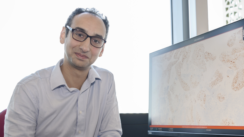 John Tadross sitting at a desk with a window behind him. On the computer screen is a pathology image from a microscope showing stained cells.