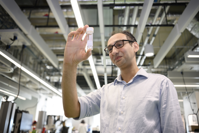 Dr John Tadross standing in a large laboratory space holding up a small clear screwtop tube containing a clear liquid.