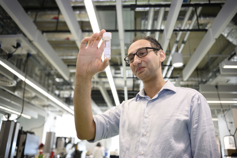 Dr John Tadross standing in a large laboratory space holding up a small clear screwtop tube containing a clear liquid.