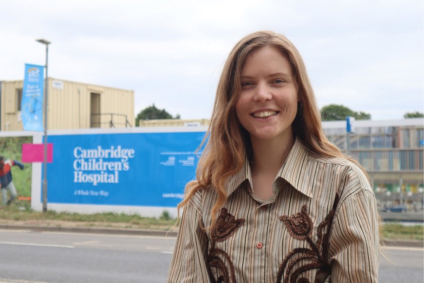 A young woman with long golden brown hair and a patterned beige shirt standing in front of the bright blue Cambridge Children's Hospital hoardings. She is smiling