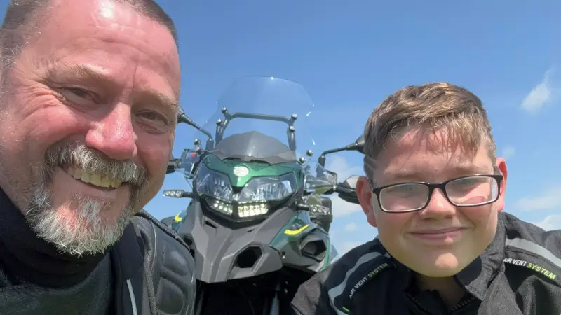 A selfie of Ken (left) and his son (right) both in black cycling gear smiling into the camera in front of Ken's dirt bike.