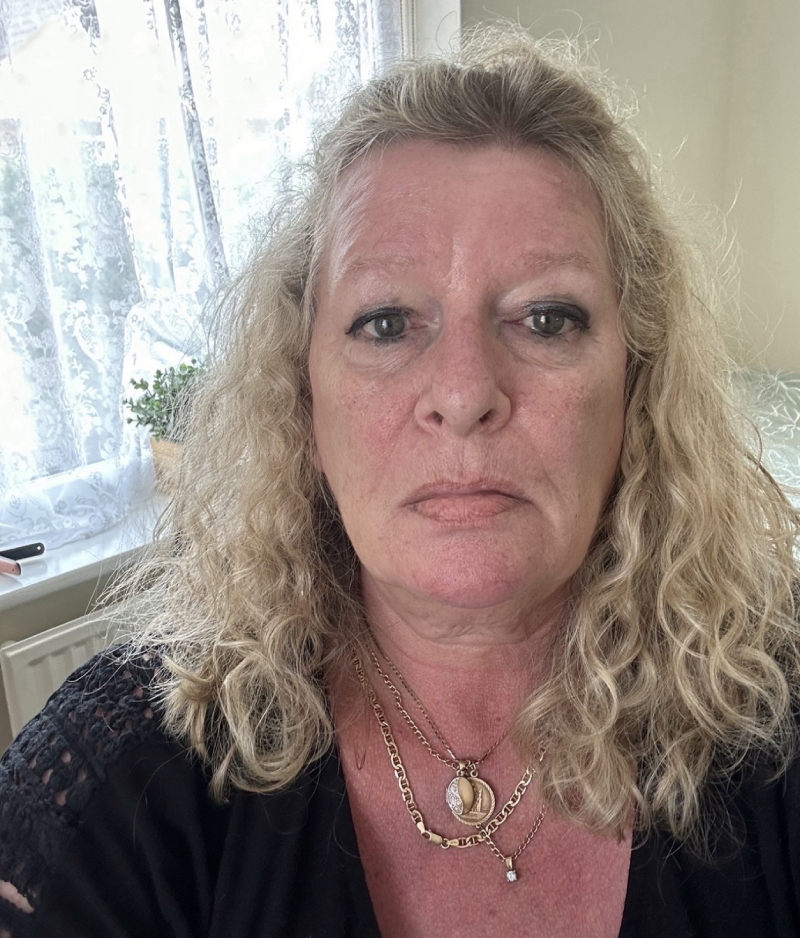Laraine facing the camera standing in front of a window in her home wearing a dark top and several necklaces