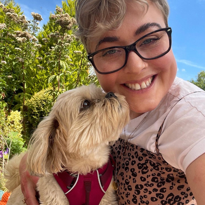 Laura, a young woman with glasses and short blonde hair smiling into the camera wearing an animal print top and holding a small dog.
