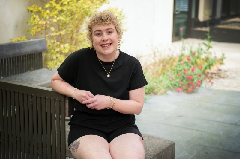 Laura, a young woman with short blonde curls sitting on a bench wearing black t-shirt and shorts