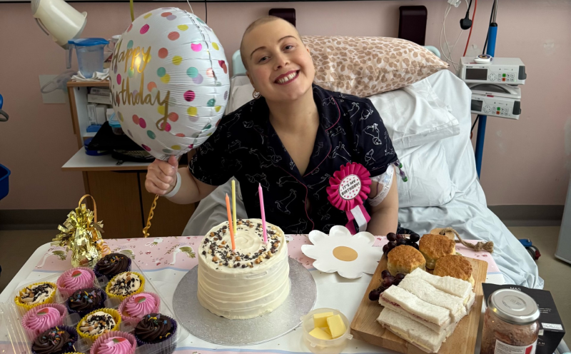 Laura sitting in a hospital bed in a black top wearing a birthday rosette and holding a balloon. A table in front of her is full of cakes and party food.