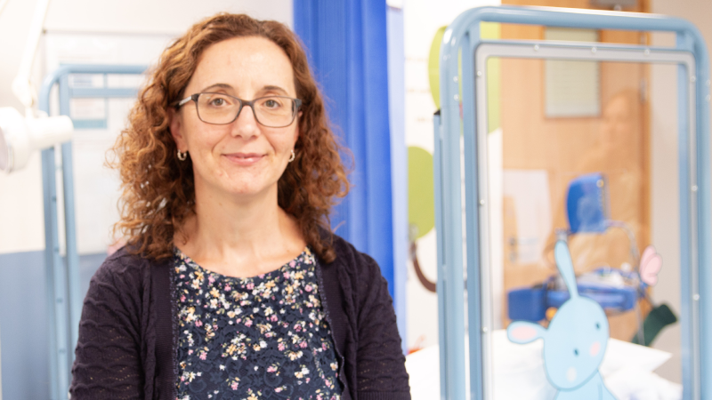 Dr Marcovecchio smiling into the camera in a blue floral top and navy cardigan sitting in front of a hospital cot.