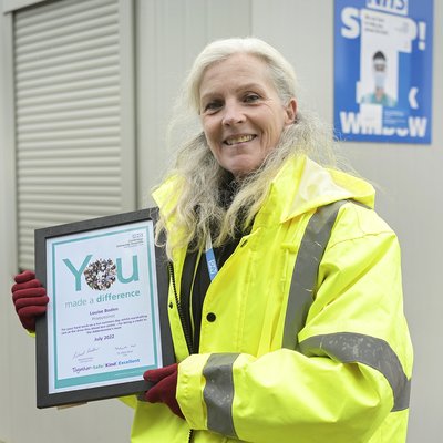 Louise has long grey hair and is stood outside at the phlebotomy drive-through. She is wearing a high-vis jacket and red gloves. She is smiling and holding her you made a difference certificate