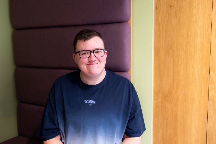 A boy wearing glasses smiling, with a purple seat behind him