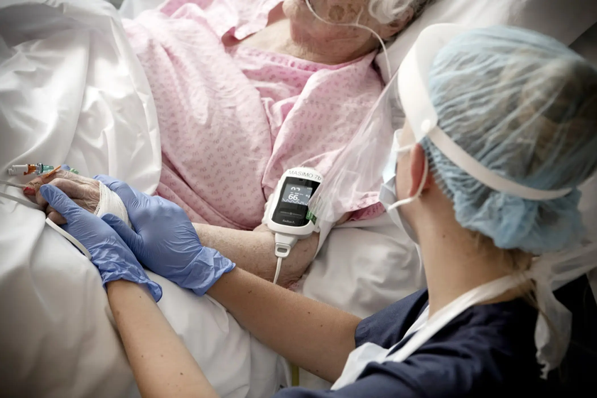 Close-up of a patient lying in a hospital bed wearing a pink patterned gown, with a pulse oximeter clipped to the patient’s finger and an IV line in place. A healthcare worker wearing blue gloves and a surgical cap gently holds the patient’s hand