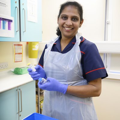 Manjumol wears a dark blue nurse's uniform with red trim, as well as blue surgical gloves and an apron. She is holding some empty blood collection vials.
