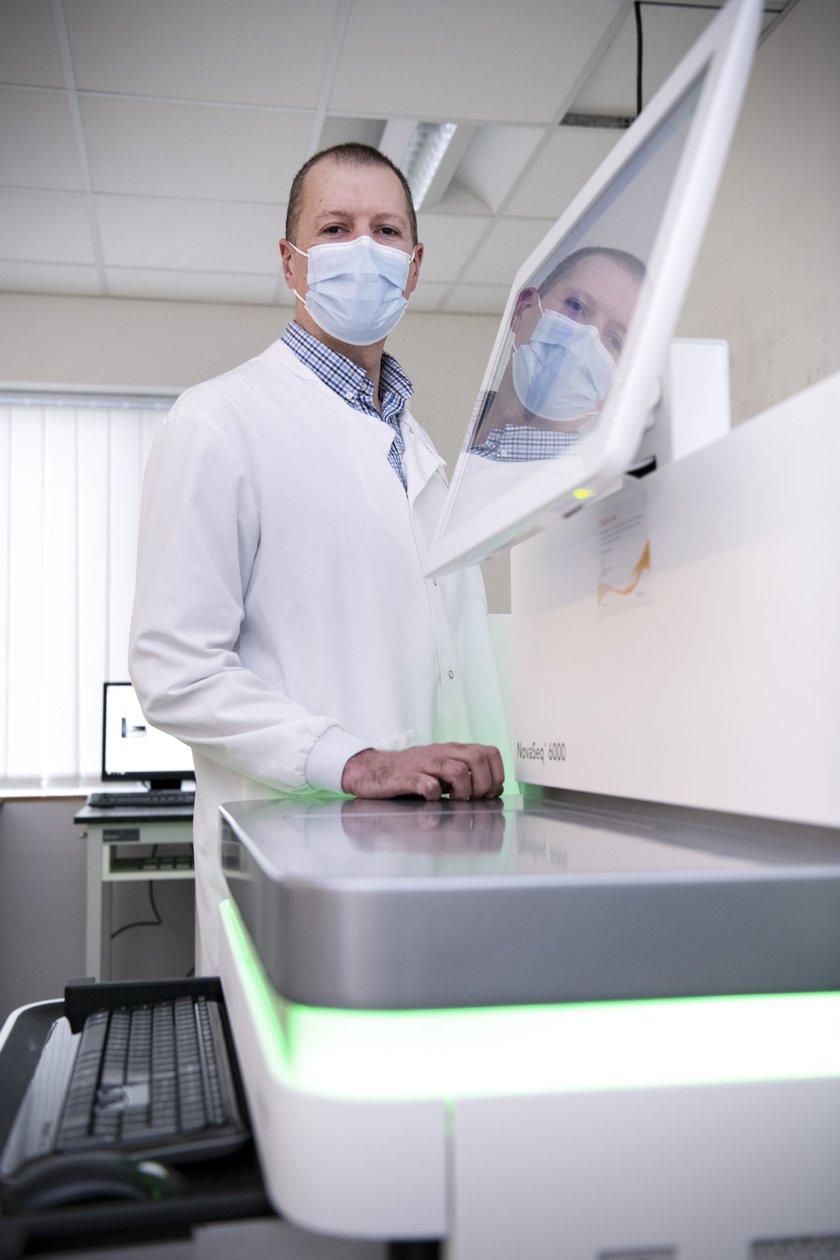 Matthew is wearing a white lab coat and a surgical mask and is stood next to a genomics laboratory machine