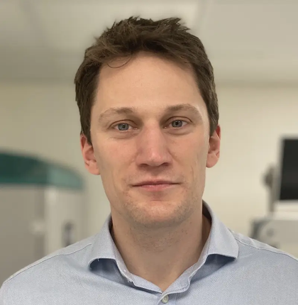 Matt facing the camera in a blue button down shirt with several machines out of focus in the background