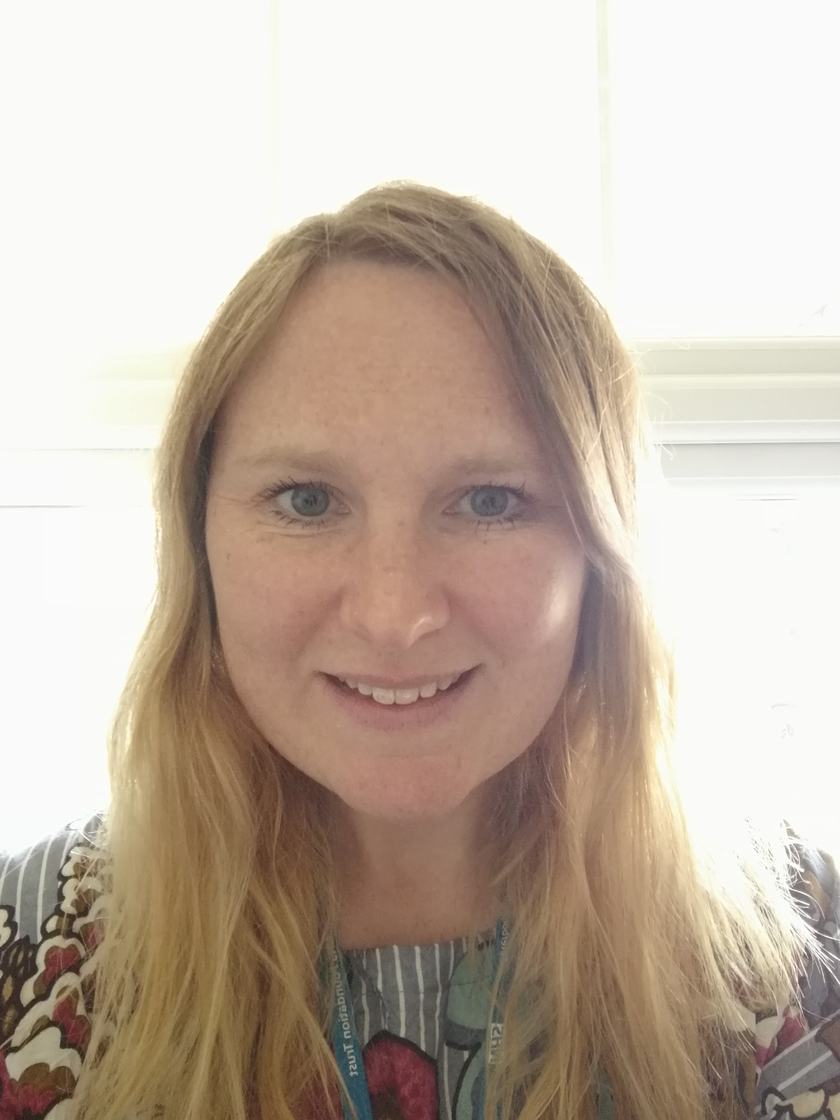 A woman with long light brown hair and a patterned top. She is smiling and looks very kind