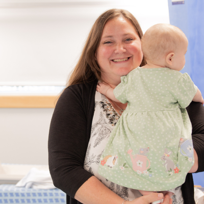 Nicole a woman with long hair in a white patterned top and black cardigan looking into the camera and holding baby Elizabeth who is facing away from the camera and wearing a green spotted dress with animals around the hem.