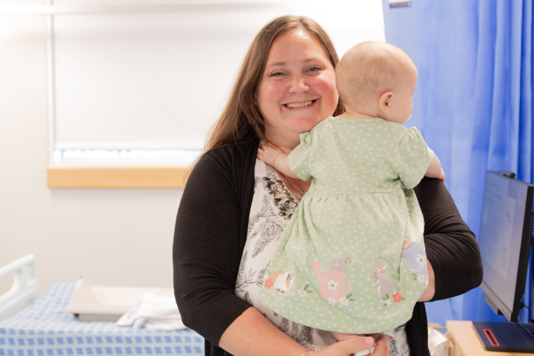 Nicole a woman with long hair in a white patterned top and black cardigan looking into the camera and holding baby Elizabeth who is facing away from the camera and wearing a green spotted dress with animals around the hem.