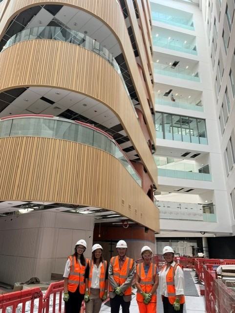 Project team are pictured standing in the Atrium at Moorfields Eye Hospital in London, it is still under construction, they are wearing high-vis and hard hats and smiling at the Camera.