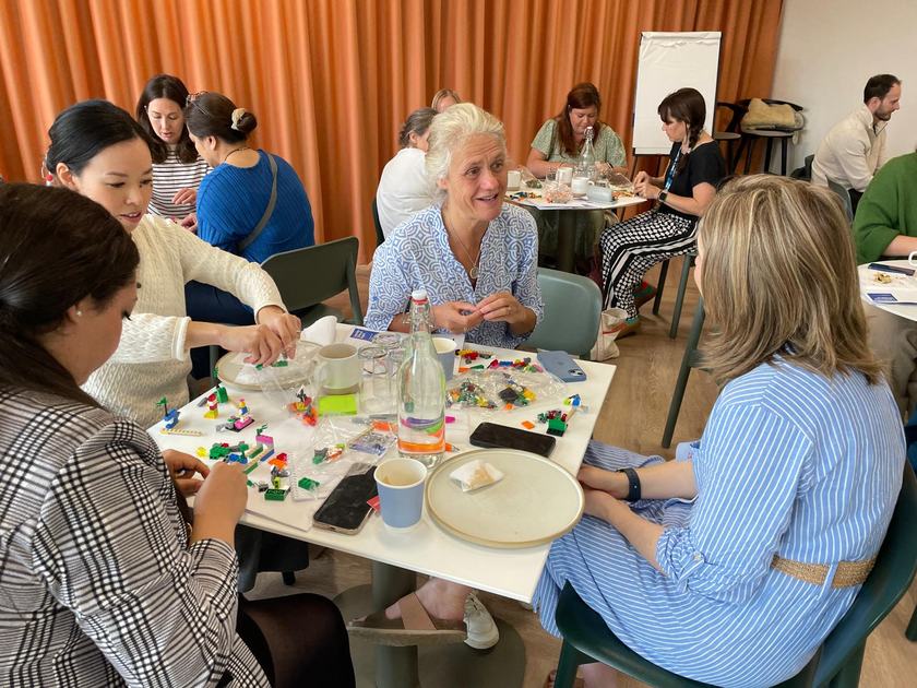 Three women at a table in a friendly work space. They are talking and writing things down. There are lego creations on the table that they've made