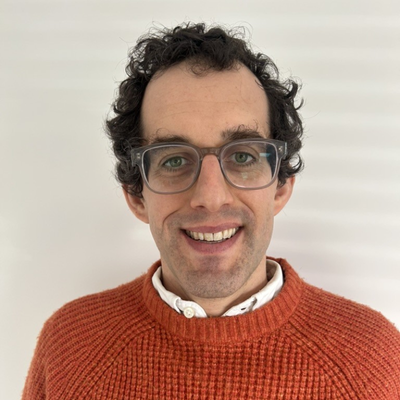Peter Hartley smiling into the camera in front of a plain background wearing an orange jumper.