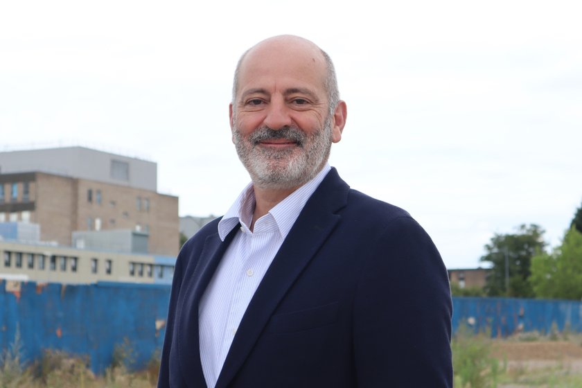 A man with dark grey hair on the sides and a dark grey beard, wearing a dark blue suit and stripy shirt. He is standing on the Cambridge children's Hospital future building site