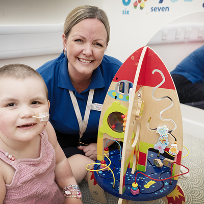 Rachel sitting on the floor with a 1 to 2 year old baby who has a feeding tube, they are both smiling looking at a toy