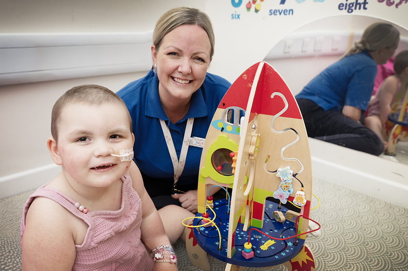 Rachel sitting on the floor with a 1 to 2 year old baby who has a feeding tube, they are both smiling looking at a toy