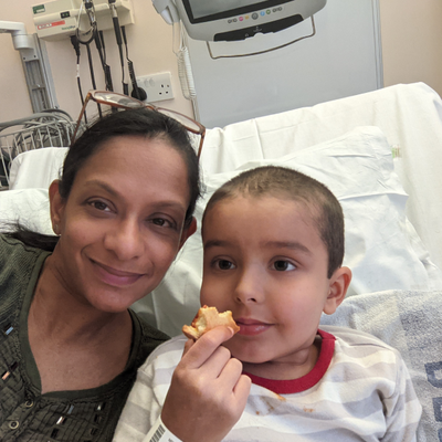 A selfie of Razia sitting on a hospital bed next to Zayan who is eating.