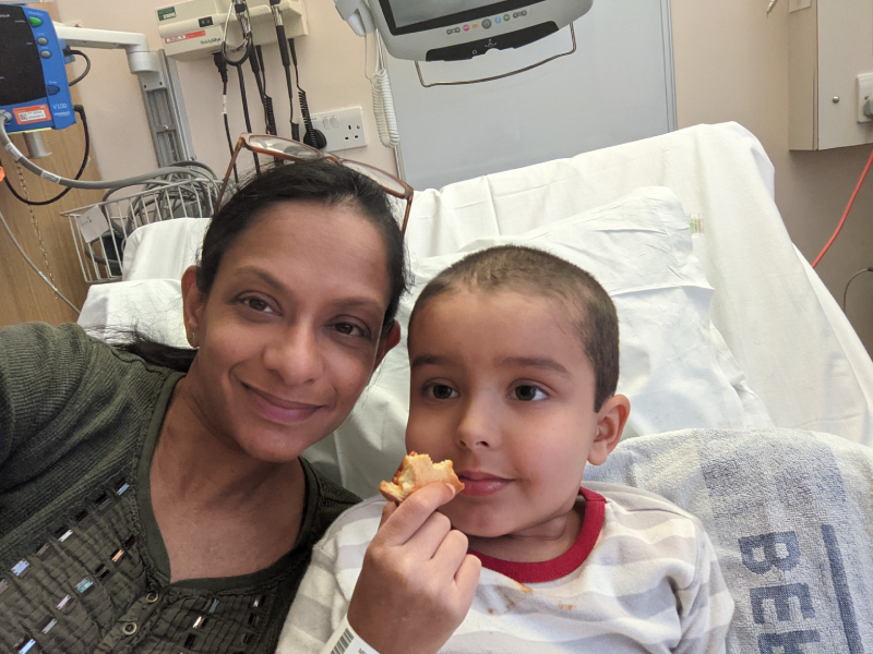 A selfie of Razia sitting on a hospital bed next to Zayan who is eating.