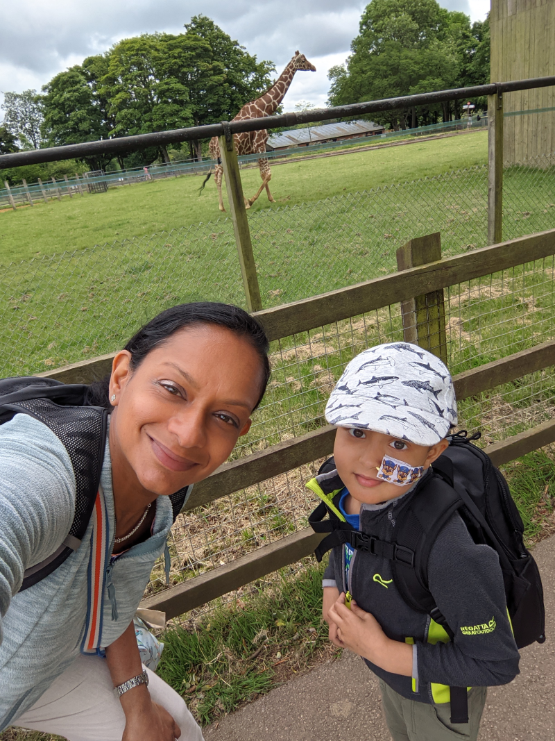A selfie of Razia with Zayan standing outside in front of a fence with a giraffe in the background.
