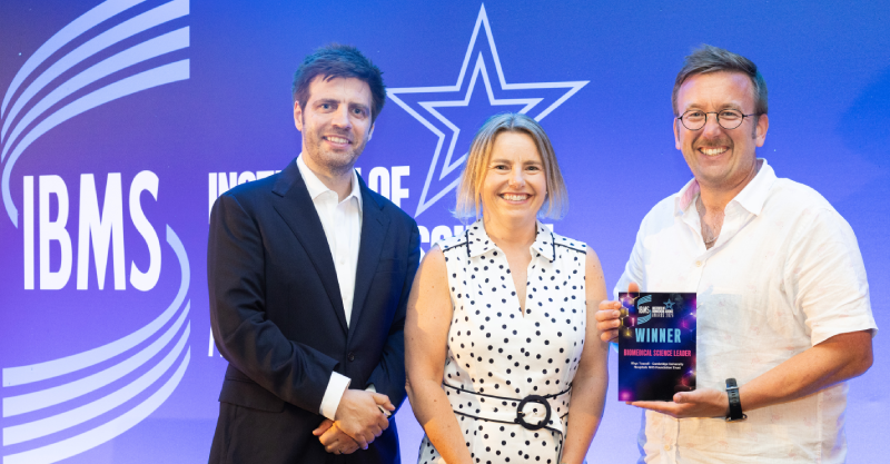 Rhys on the right with his award next to the presenters of the award all standing in front of a blue IBMS background.
