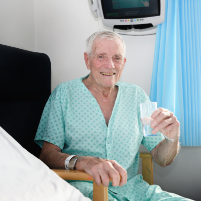 A NHS male patient sitting on a hospital chair, wearing a green hospital gown and smiling