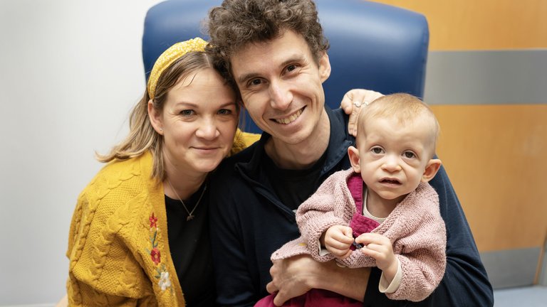 A woman with a yellow cardigan, yellow hairband and blond hair, sitting next to a man with dark curly hair. He is holding a baby on his lap. She is wearing a pink top and dark pink dungarees