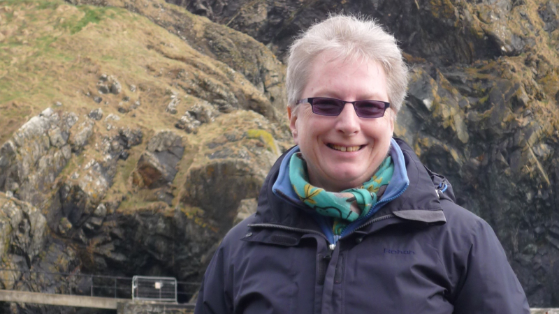 Sally smiling into the camera wearing sunglasses, a black coat and colourful scarf in front of rugged coastal cliffs