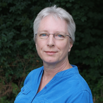 Sally side on with her head turned to face the camera wearing a powder blue button down blouse with grass and trees in the background