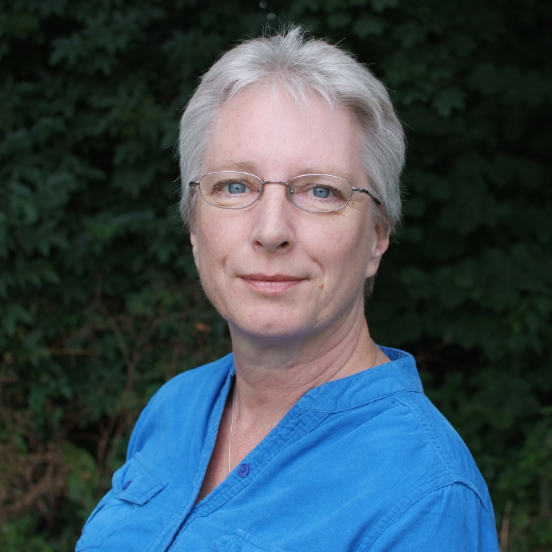 Sally side on with her head turned to face the camera wearing a powder blue button down blouse with grass and trees in the background