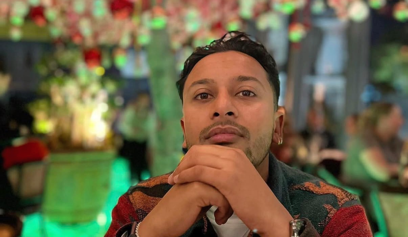 Sameer wearing a patterned shirt looking towards the camera sitting in a restaurant with a tree of lights in the background