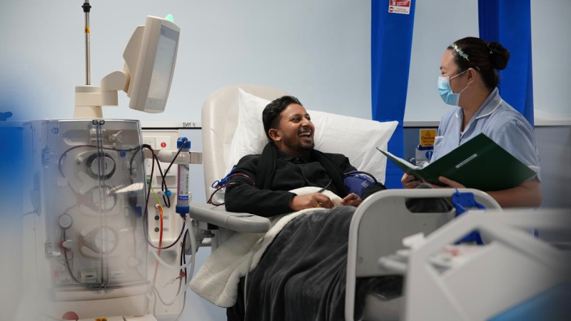 Sameer sitting connected to a haemodialysis machine laughing with a member of NHS staff