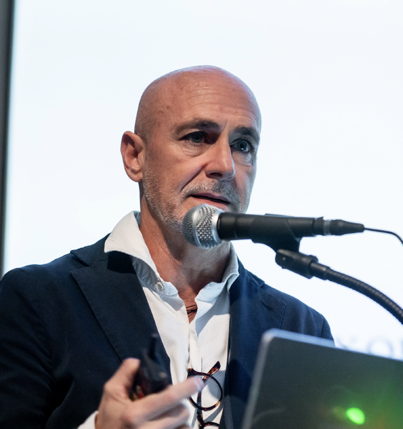 Stefano Pluchino presenting at a conference in front of a projector screen with a microphone in the foreground