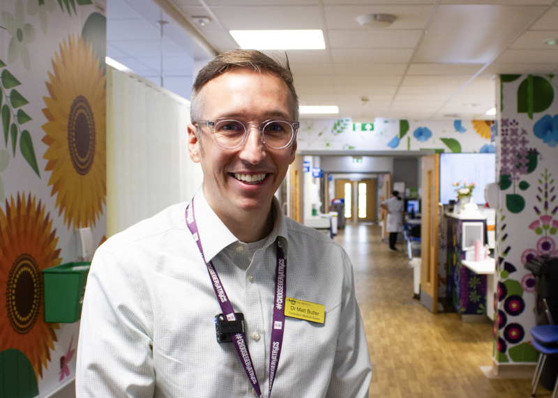 Dr Butler standing in the corridor of T2 in a white button down shirt. Behind him are many large geometric floral prints covering the walls around the ward.