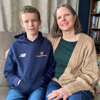 A teenage boy with short fair curly hair, blue eyes and braces, wearing a blue hoodie. His mum has brown bobbed hair and is wearing a brown cardigan and dark green top