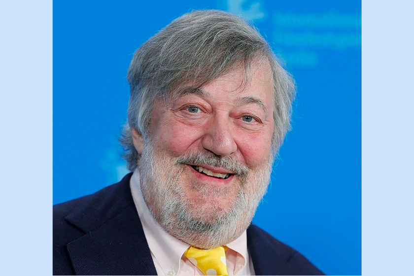A man with grey floppy hair and grey beard, with a yellow spotty tie, white shirt and smart black jacket