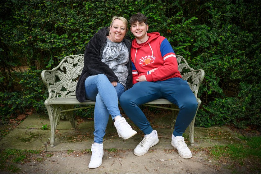 A woman with short blond hair and a black fleece jacket and jeans sitting on a white patterned bench with a teenage boy who has brown tufty hair and a red and blue hoodie