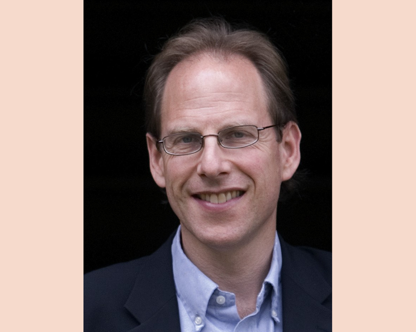 A man with brown slightly receding hair and rectangular rimmed glasses. He is wearing a pale blue shirt and suit jacket