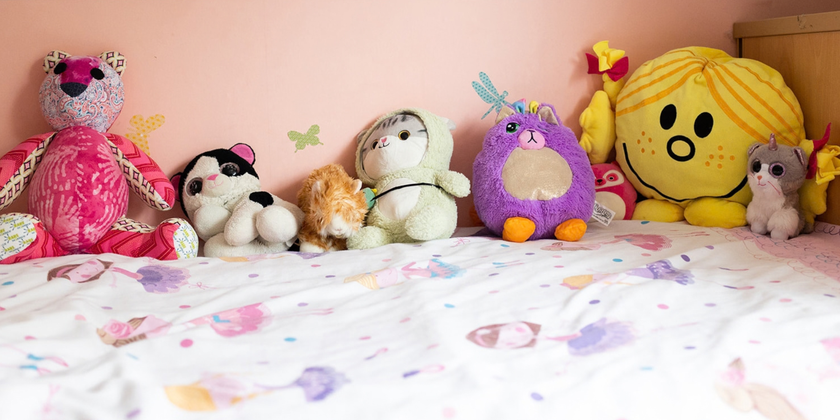 A row of colourful soft toys lined up on a child's bed with a pink wall behind