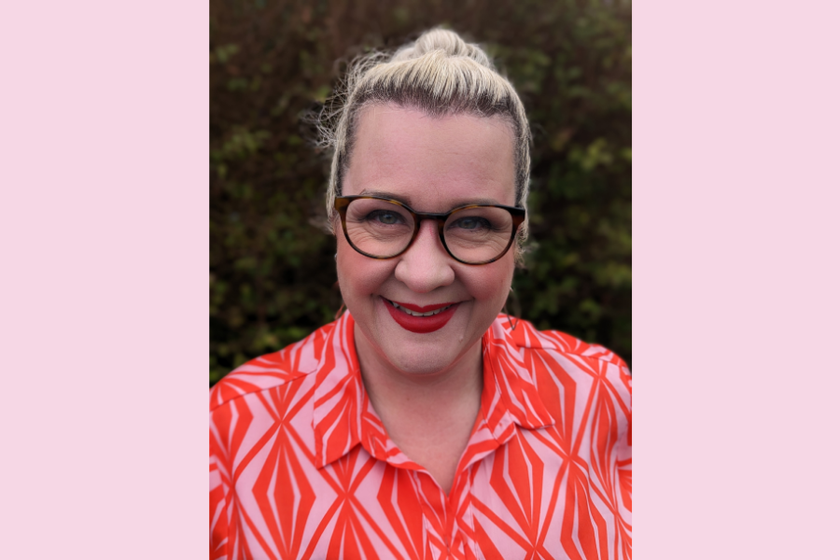 A woman with a pink and orange shirt, glasses, dark red lipstick and blond hair in a top knot. She is smiling broadly