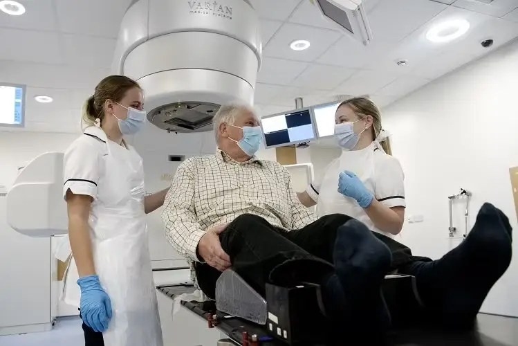 A patient wearing a mask sat up on a radiotherapy scanner. Two radiographers, wearing surgical gloves and masks are stood either side of the patient.