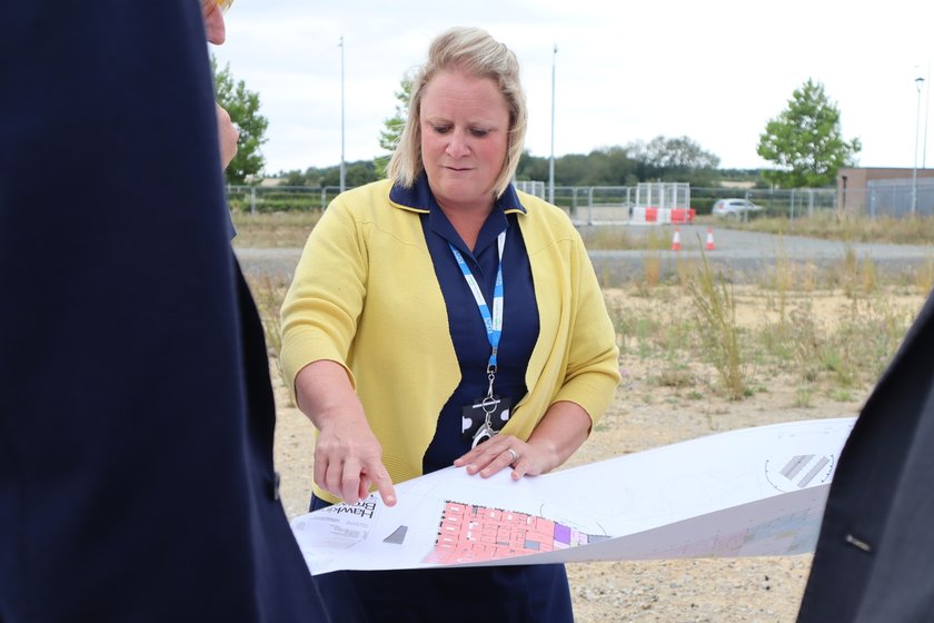 A woman with blond hair, a navy nurse uniform and bright yellow cardigan, standing on a the children's hospital future building site, pointing to a location on an architect floorplan. She is showing this to people standing around her