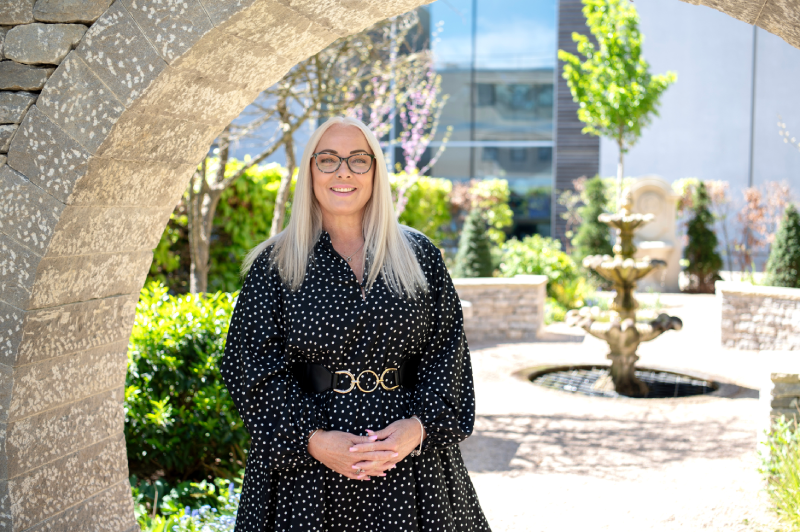 Victoria in a black spotted dress standing in front of a moongate in a sunny garden